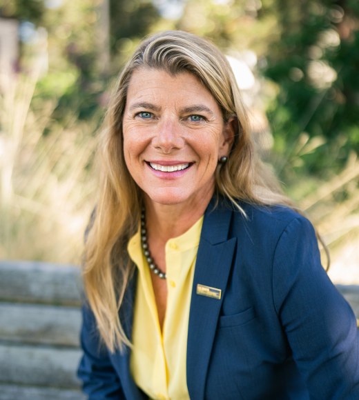 Headshot photograph of smiling blonde woman in navy blazer and yellow blouse, outdoor background