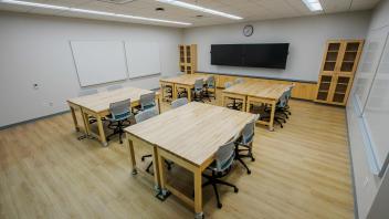 Empty modern classroom with wooden worktables, rolling chairs, whiteboards, and a wall monitor