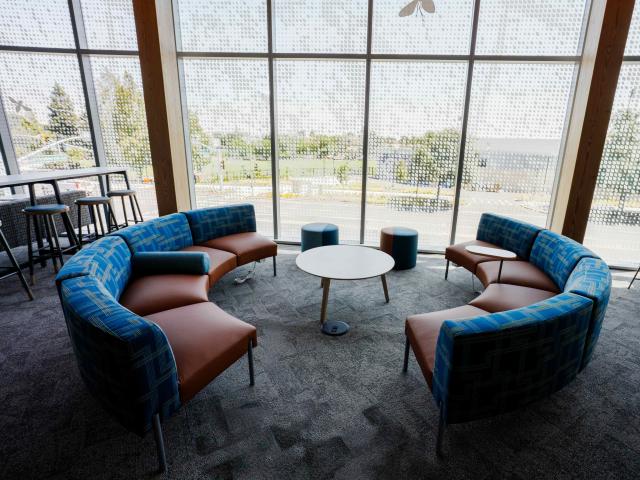 Curved blue-and-tan sofas around round tables by floor-to-ceiling windows overlooking trees.