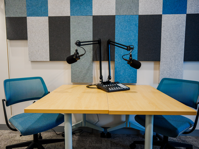 Recording table with two boom microphones, mixer, blue chairs, and acoustic panels