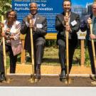 Five officials smiling with gold shovels on a raised platform at a groundbreaking